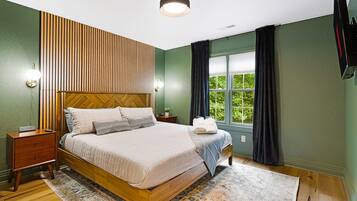Une chambre avec un lit en bois, une table de chevet, une lampe fixée au mur, une fenêtre avec des rideaux et une vue sur la verdure à l’extérieur.