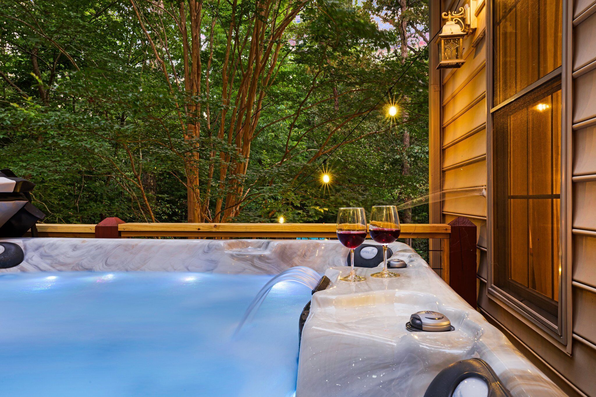 Un bain à remous avec une terrasse en bois, entouré d’arbres et d’un bâtiment en bois doté d’une fenêtre.