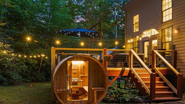 Une structure en forme de tonneau, faite de bois, avec un escalier menant à une terrasse en bois, le tout entouré de guirlandes lumineuses et de verdure.