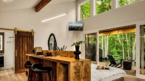 A modern bedroom with a wooden bed, a desk, and bar stools. There is a large window with a view of trees. - The 110 Copse (Wartburg)
