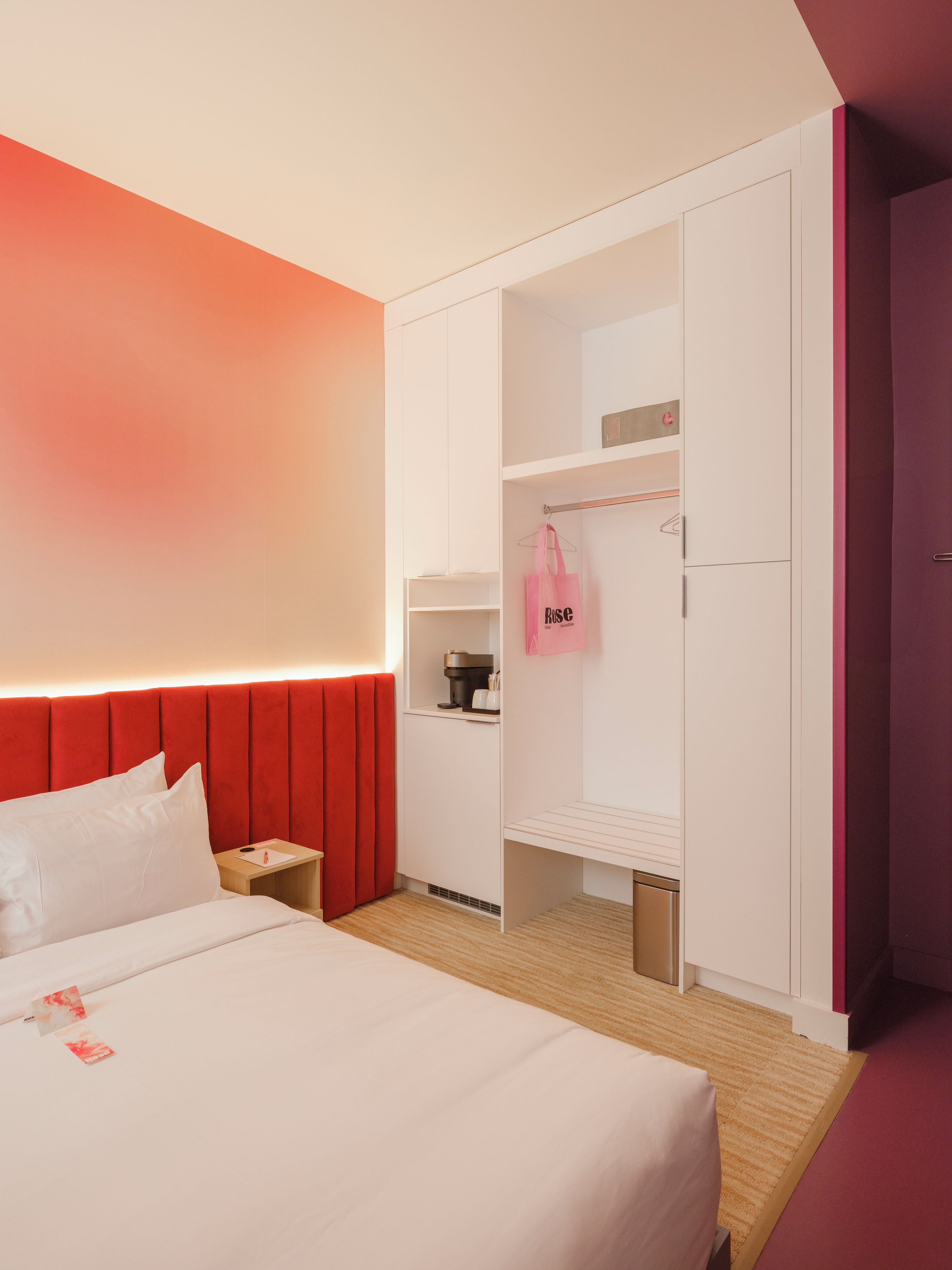 A hotel room with a bed, a red headboard, a wardrobe, and a small kitchenette area.