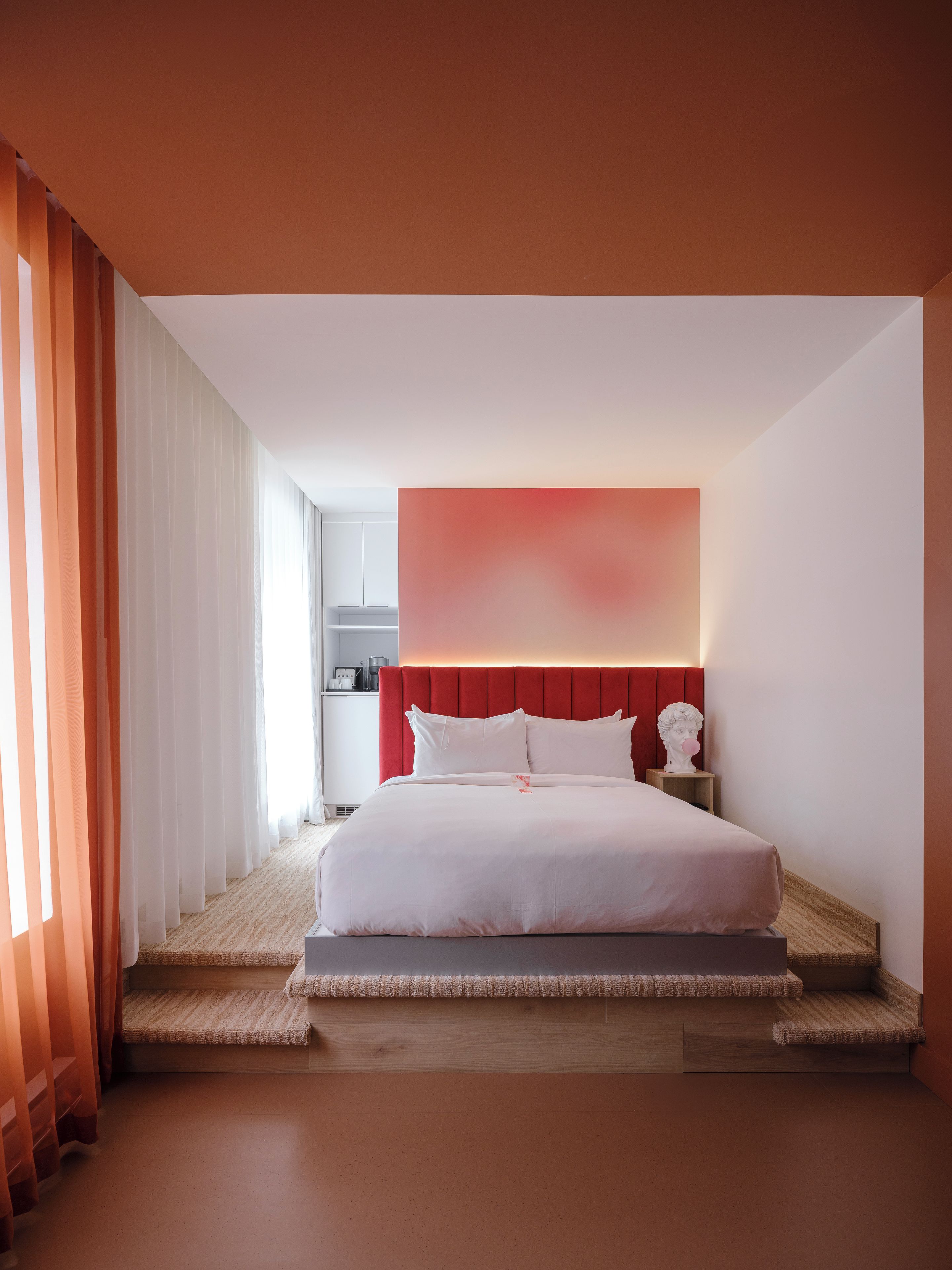 A modern hotel room with a large bed, a red accent wall, and a view of the adjacent kitchen.