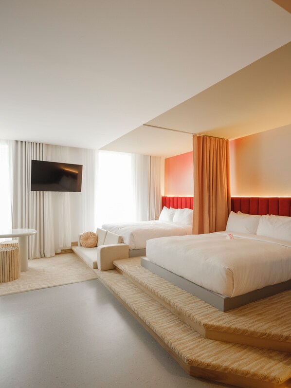 A hotel room with a large bed, a smaller bed, a flat-screen TV mounted on the wall, a seating area with a table, and a window with curtains.