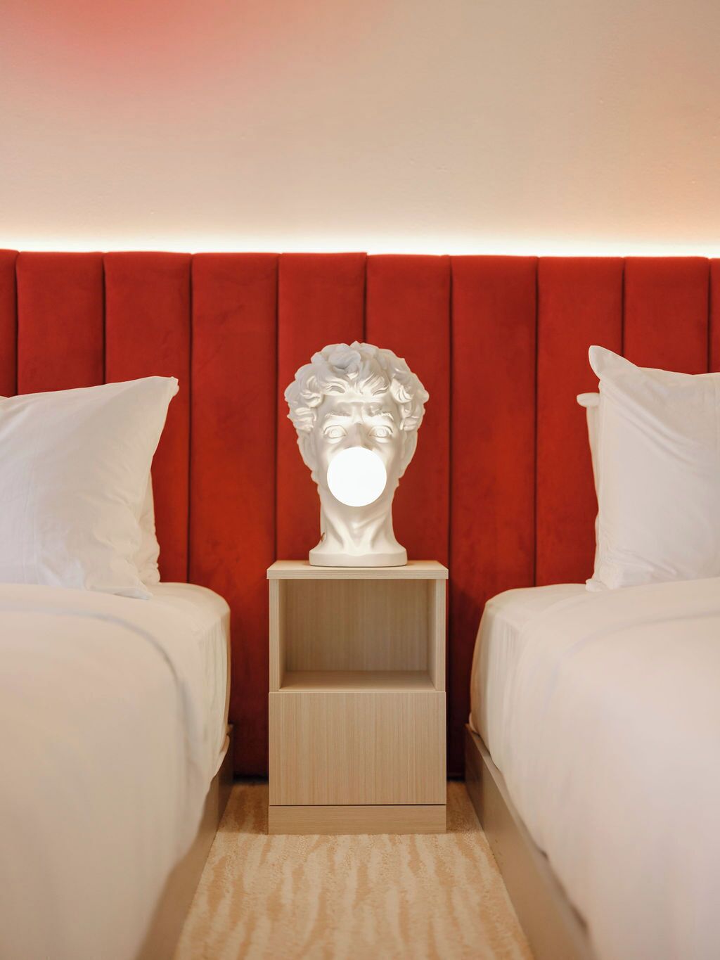 Habitación de hotel con dos camas, cabecera roja, escultura de busto blanco sobre una mesita de noche y un espejo iluminado.