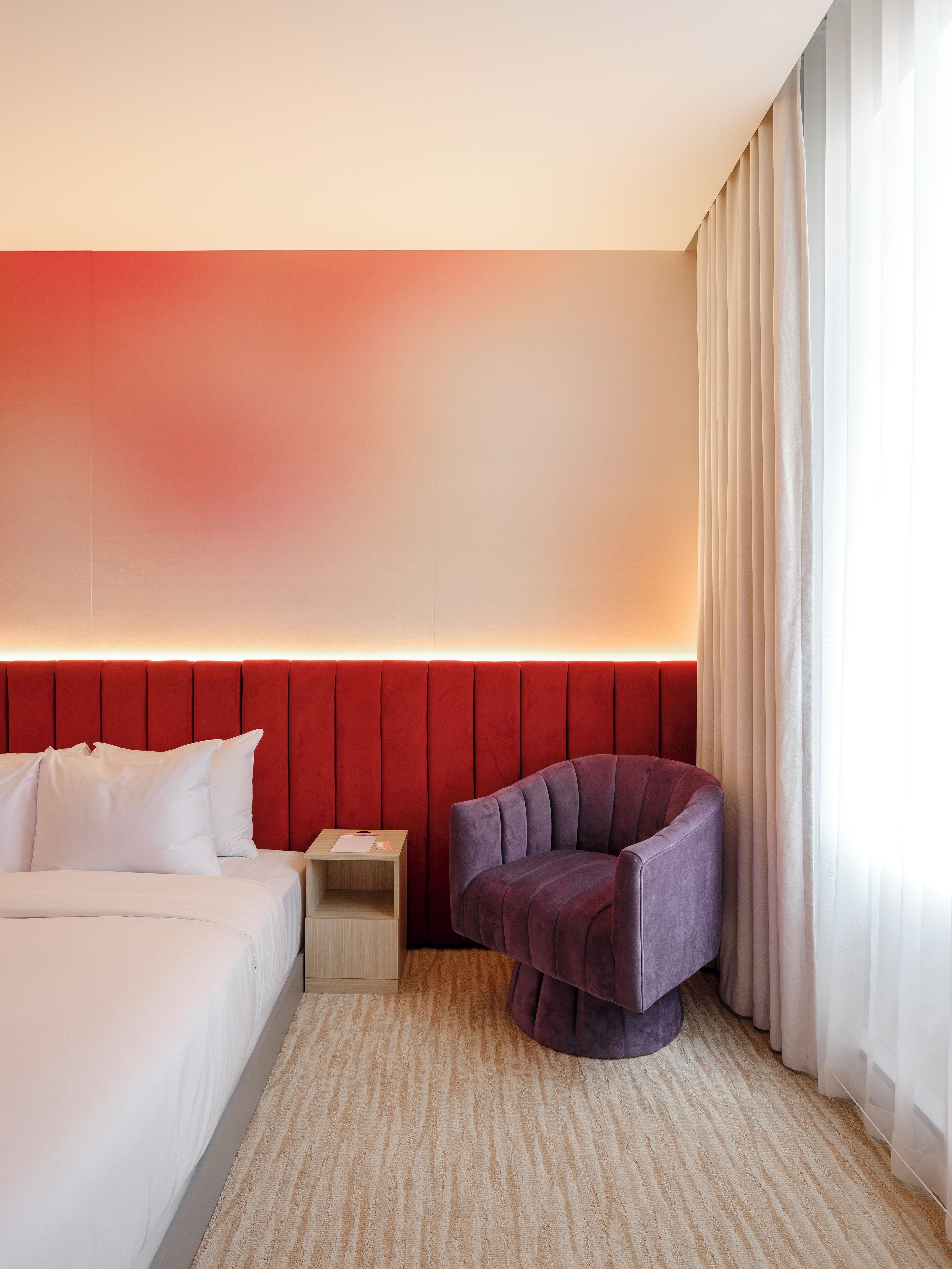 A hotel room with a bed, a nightstand, a chair, and a red headboard.