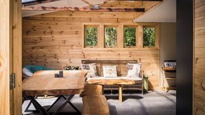Un interior de madera acogedor con sofá, mesa y sillas, una ventana con vistas a la vegetación y un techo con vigas de madera.
