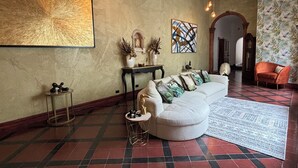A living room with a large abstract painting, a white sofa with patterned cushions, a small round table with a gold base, and a patterned rug. - The Victorian Hotel CR (San José)