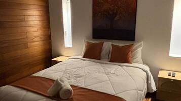 A bedroom with a wooden headboard, a large bed with a quilt, two pillows, a blanket, and a nightstand with a lamp.