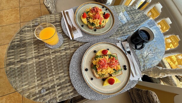 A breakfast setup with two plates of scrambled eggs and toast, a glass of orange juice, and a cup of coffee on a wicker table. - The Victorian Hotel CR (San José)