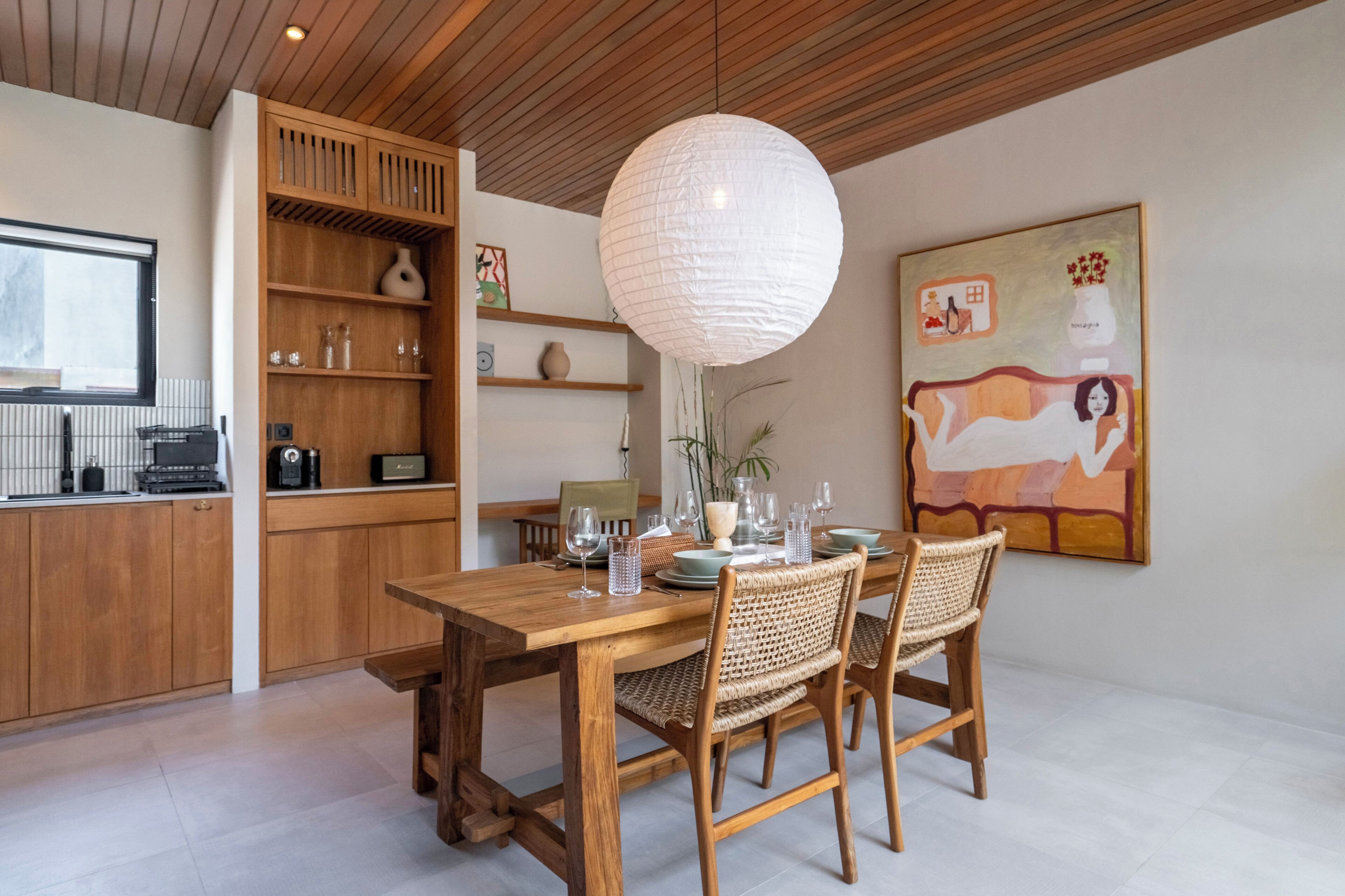 A dining area with a wooden table and chairs, a hanging light fixture, and a painting on the wall.