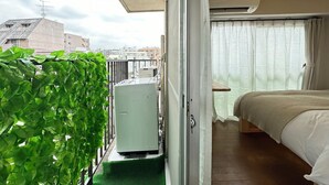 Ein Balkon mit einer Zimmerpflanze, einem kleinen Tisch und einem Bett mit weißer Bettdecke.