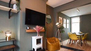 Una sala de estar con televisor, chimenea, zona de comedor con sillas amarillas y un ventanal grande.