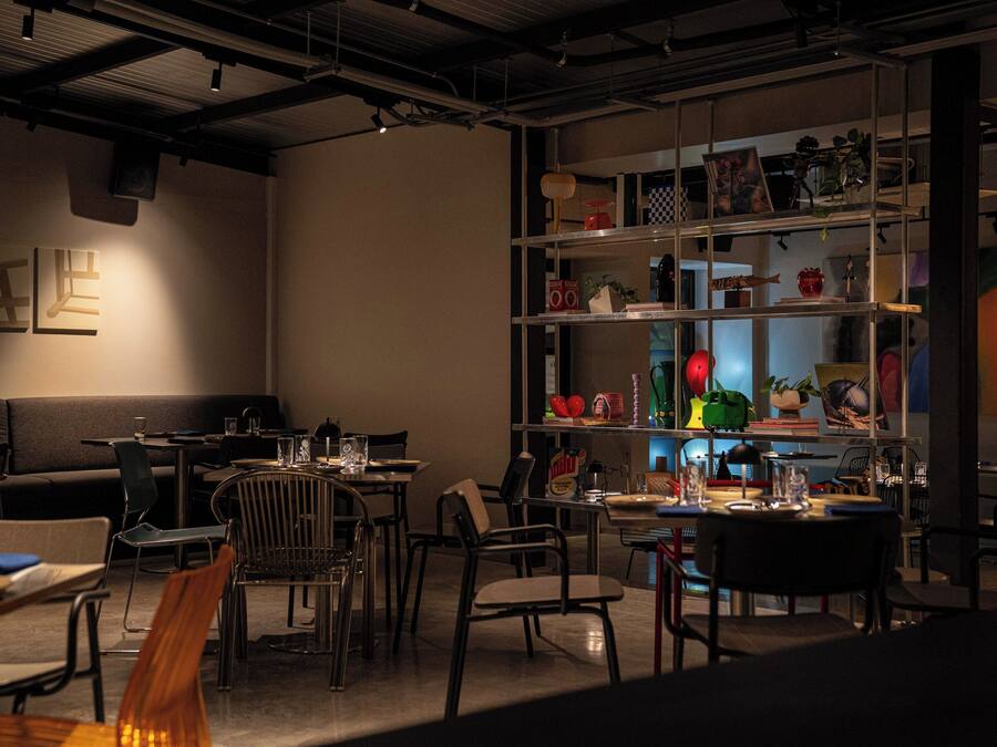 Un comedor moderno con mesas y sillas, una estantería con diversos objetos, y un sofá al fondo.