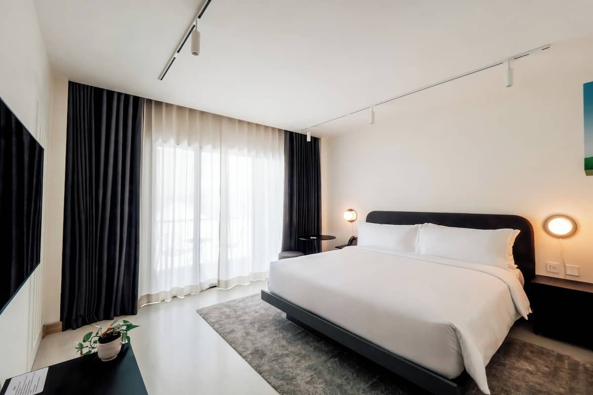 Ein modernes Hotelzimmer mit einem großen Bett, einem Schreibtisch mit Pflanze und Blick ins Freie durch transparente Vorhänge.