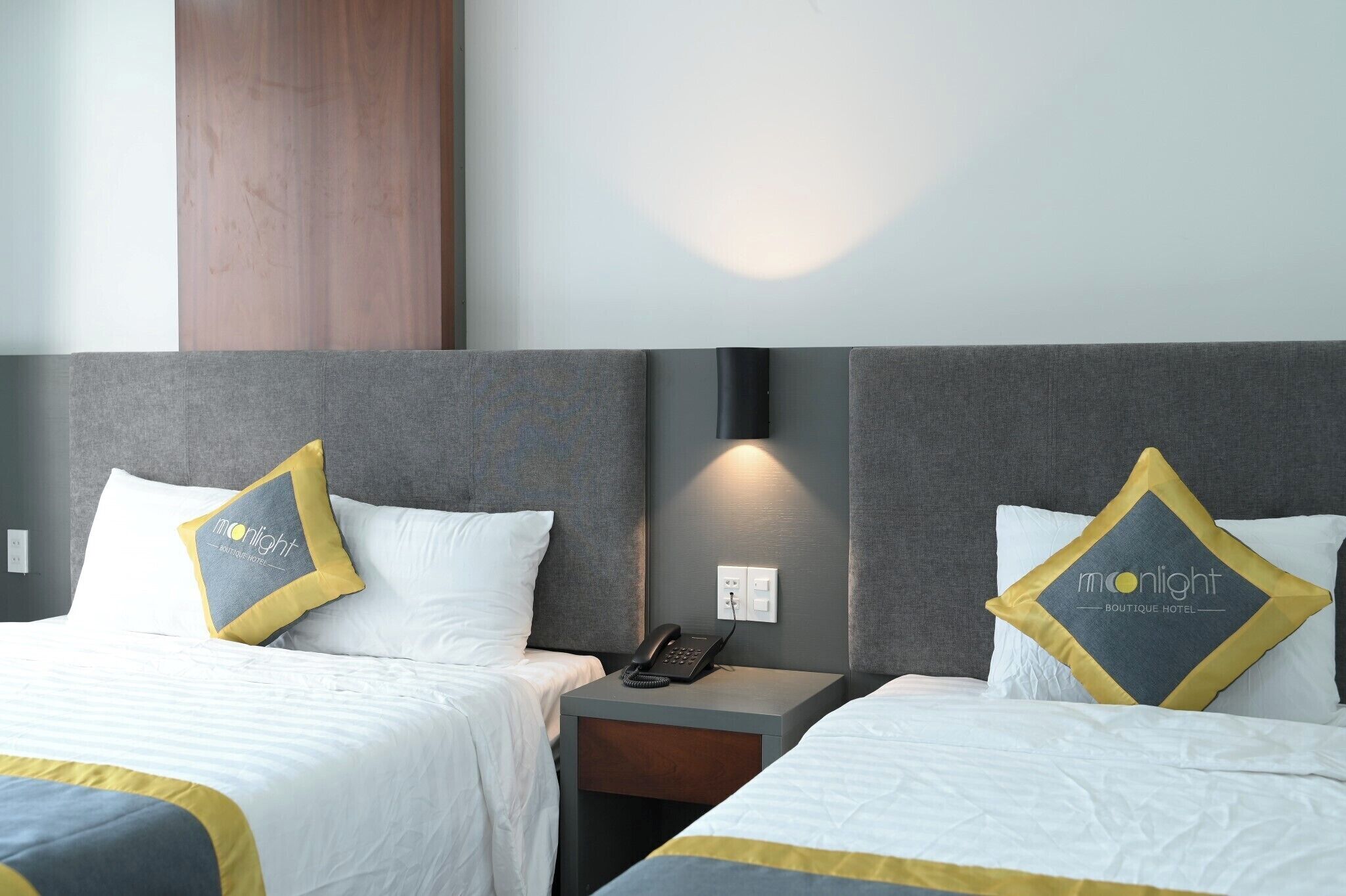A hotel room with two beds, each with a grey headboard and a white bedspread. There are two pillows with a logo on them, and a bedside table with a phone and a lamp.