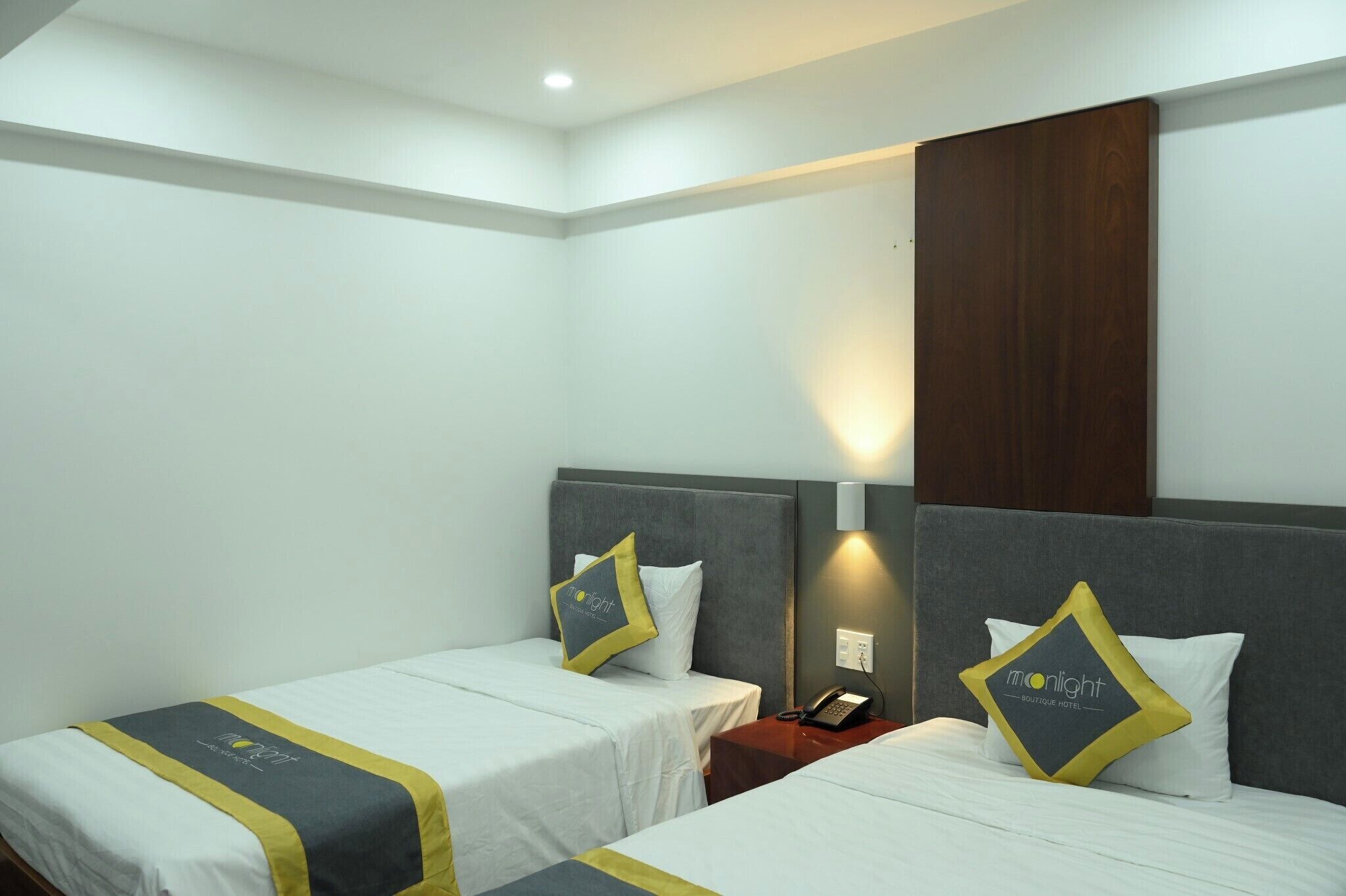 Hotelkamer met twee bedden, elk met een grijs hoofdeinde, wit beddengoed en een geel kussen. Er is een houten paneel aan de muur en een nachtkastje met een telefoon.