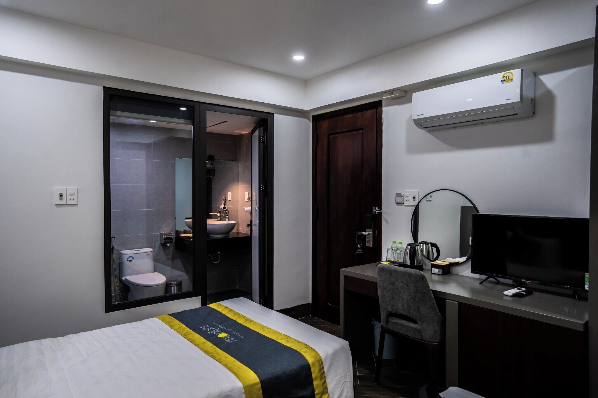 A hotel room with a bed, a desk with a computer, a TV, and a bathroom visible through a glass door.