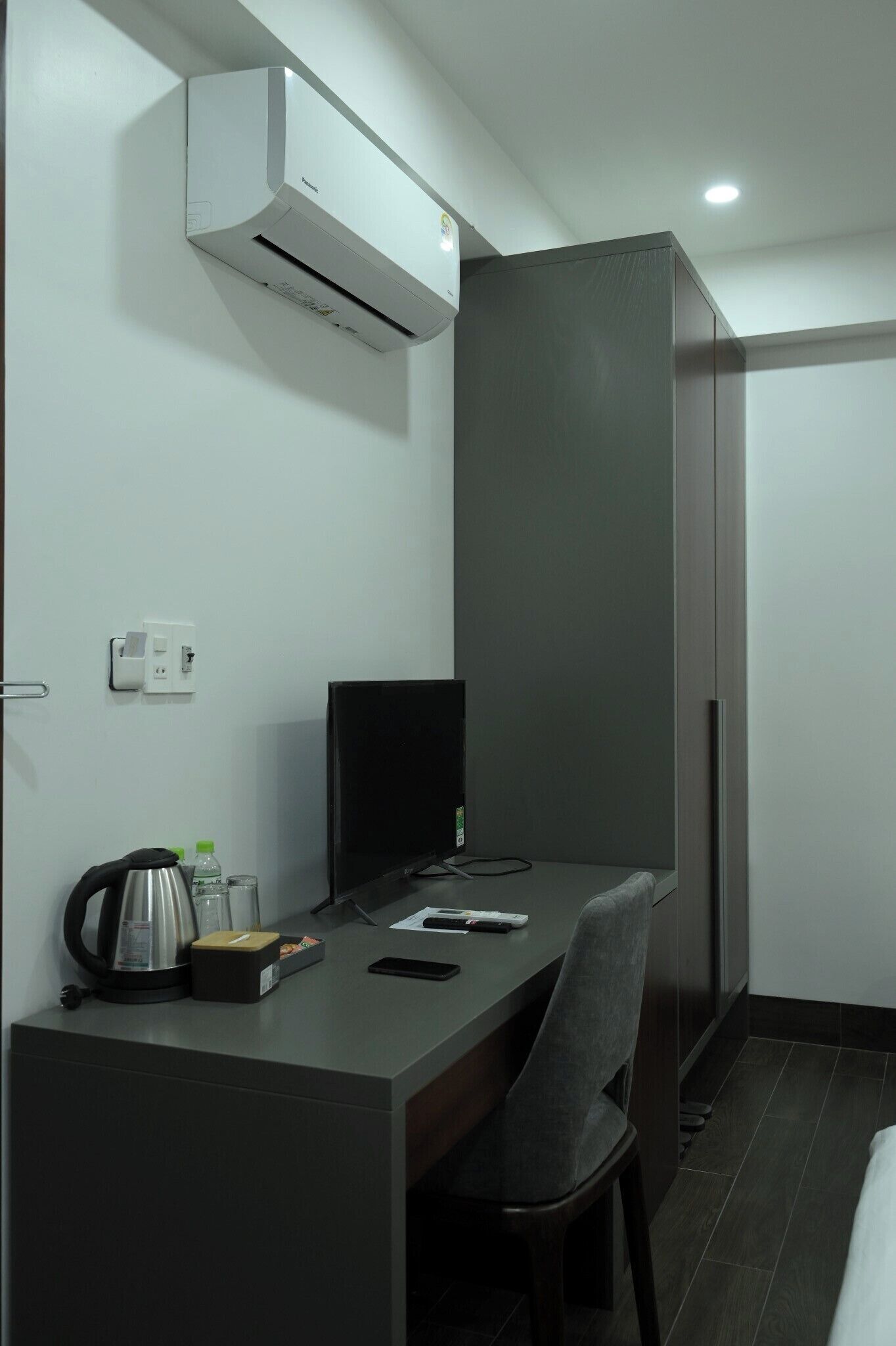 A compact hotel room with a desk, chair, and a built-in air conditioner.