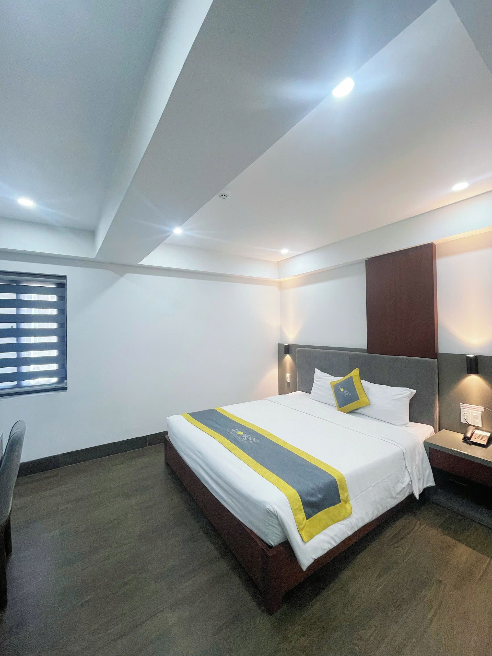 A hotel room with a bed, a desk, a chair, a window with blinds, and a wall-mounted telephone.