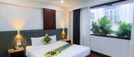 A hotel room with a bed, bedside lamps, a window with curtains, and a view of greenery outside.