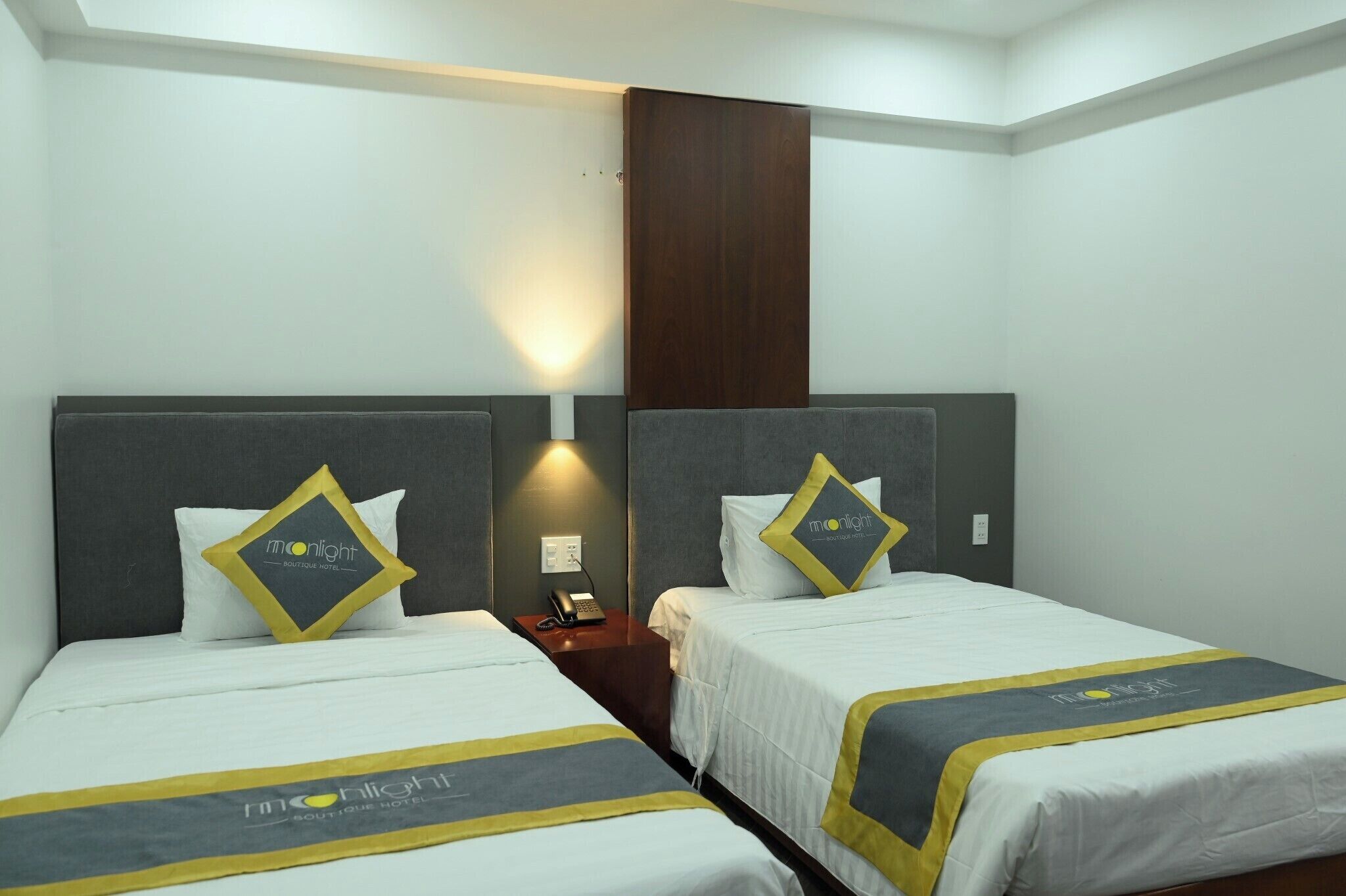 Une chambre d’hôtel avec deux lits, chacun doté d’une tête de lit grise et d’une couvre-lit blanche. On y trouve deux tables de chevet avec des lampes, ainsi qu’un panneau en bois sur le mur.