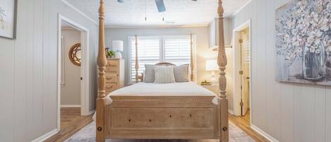 Een hemelbed met houten afwerking, een plafondventilator en een groot raam met zonwering.