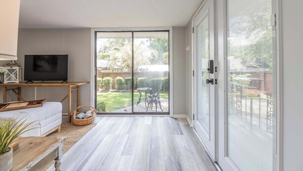 A modern living room with a flat-screen TV, a sofa, a wooden table, and a large sliding glass door leading to a garden. - Camille Cottage ~ Ideal for Short-Term Stays or Traveling Professionals (Columbus)