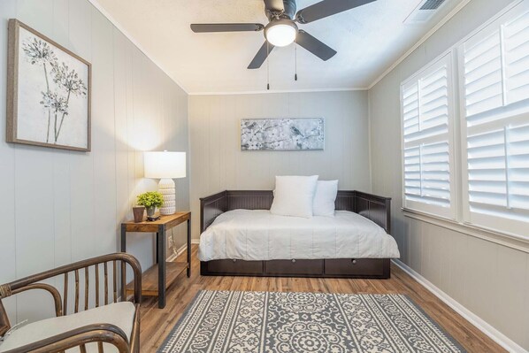 Un dormitorio con cama, ventilador de techo, mesita de noche con lámpara, una silla y una alfombra estampada.