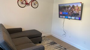 A living room with a grey sofa, a mounted TV displaying a show, and a red bicycle on the wall. - Spacious 3-bedroom Suite in fabulous Surrey Wi-Fi, EV Charging, Basketball Court (Surrey)