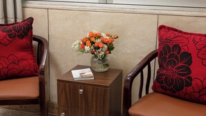 Une pièce comprenant deux chaises en bois, chacune agrémentée d’un coussin rouge orné de motifs floraux noirs. Une petite table de chevet en bois supporte un vase en verre contenant des fleurs orange et blanches, et un magazine est posé entre les deux chaises.