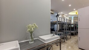 Une salle de bain moderne avec deux lavabos, un miroir et un vase rempli de fleurs blanches. On aperçoit, au fond, la structure d’un lit superposé.