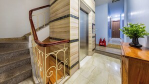 Un hall d’hôtel avec un escalier en marbre, un ascenseur et un comptoir de réception agrémenté d’une plante en pot.