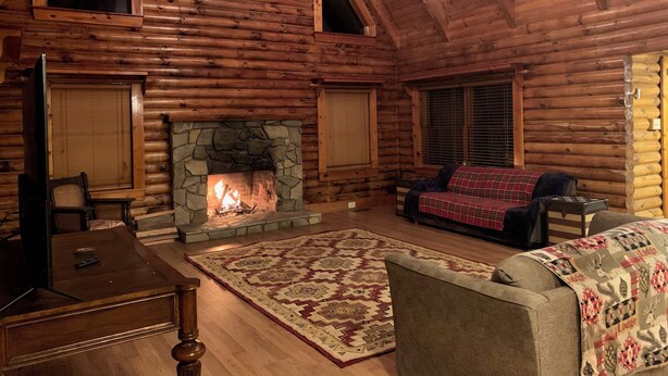 A log cabin interior with a stone fireplace, wooden furniture, and a high ceiling.