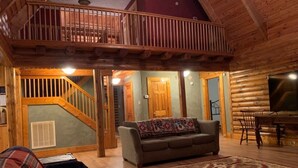 A log cabin interior with a wooden staircase, a sofa, a patterned rug, and a television.
