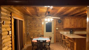A log cabin interior with a dining table, chairs, a kitchen area, and a ceiling fan.