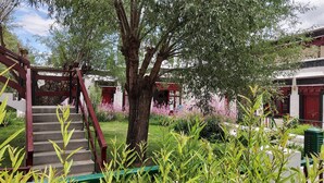 A traditional building with a staircase leading to a red-roofed structure, surrounded by lush greenery and flowering plants. - Lharimo North Cottage (Leh)