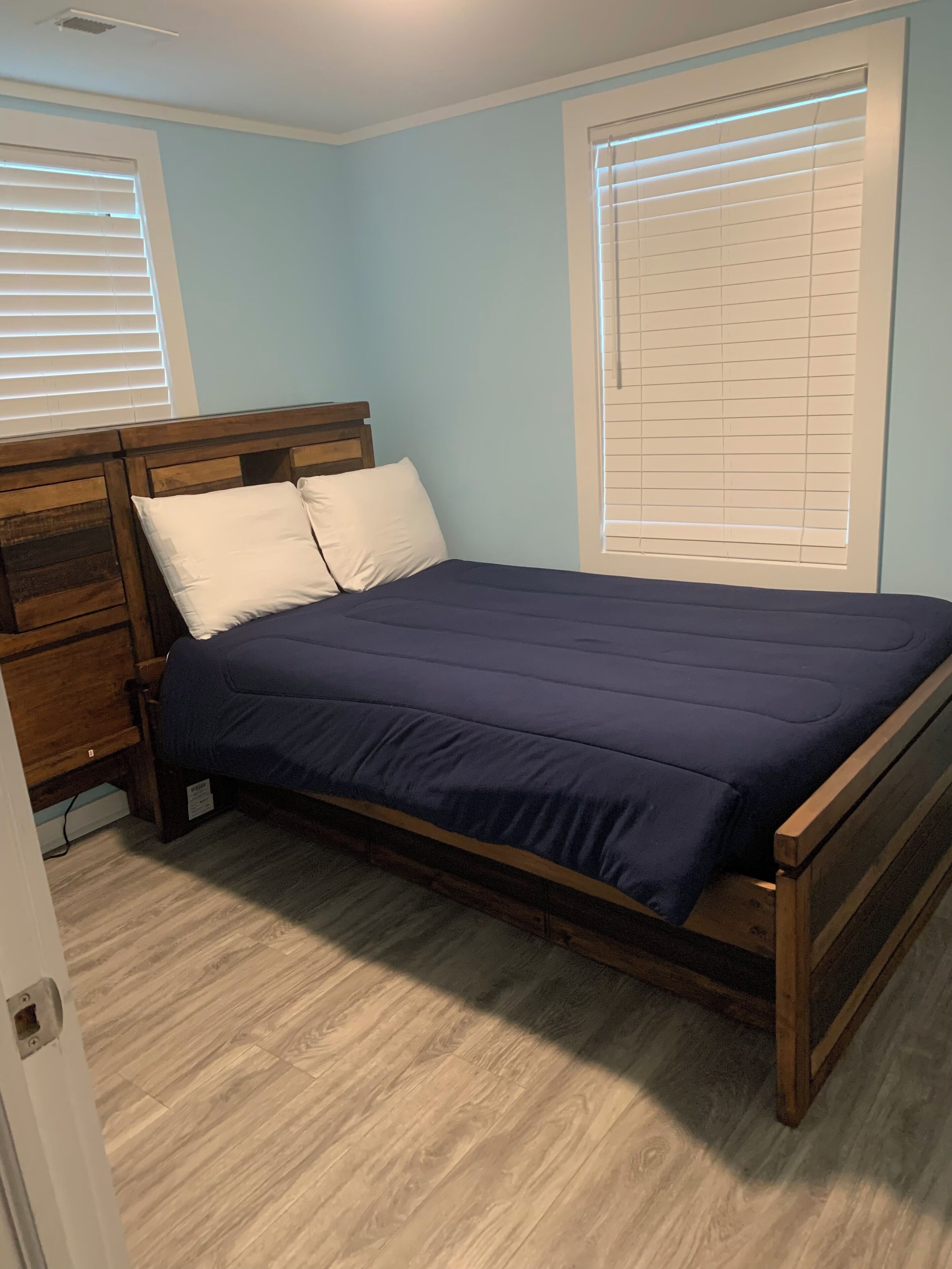 A bedroom with a wooden bed frame, a dark blue bedspread, and white pillows. There is a wooden nightstand to the left of the bed.