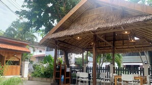 A covered outdoor seating area with a thatched roof, surrounded by greenery and a concrete floor. - La nuit de Laos (Luang Prabang)