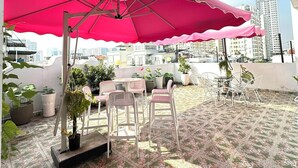 A rooftop terrace with a patterned floor, white chairs, and a large pink umbrella.