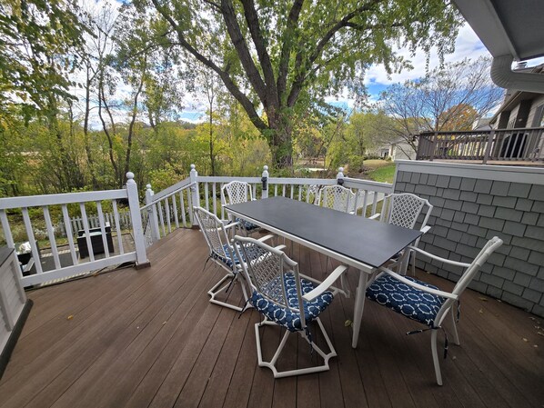 A wooden deck with a table and chairs, surrounded by a white railing, overlooking a garden and trees.