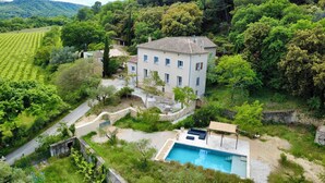 Una casa de dos pisos con piscina, rodeada de exuberante vegetación y un muro de piedra.