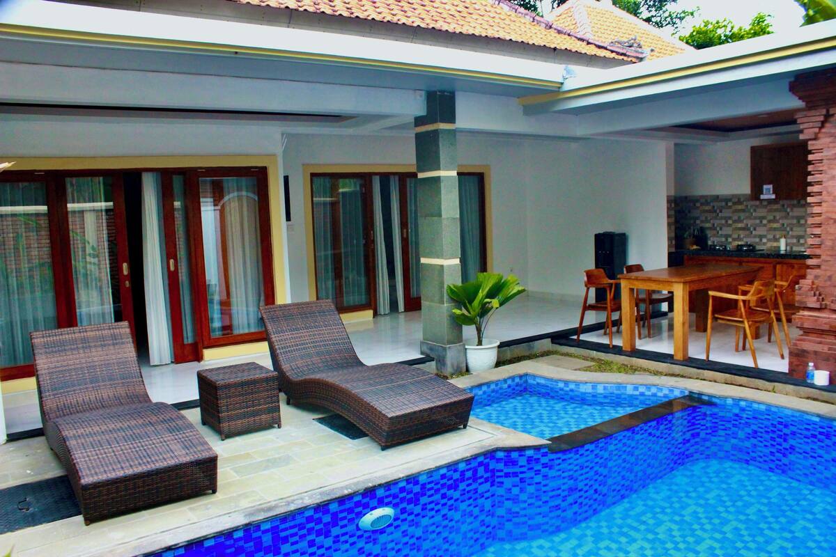 A swimming pool with lounge chairs, a dining table, and a kitchen area.
