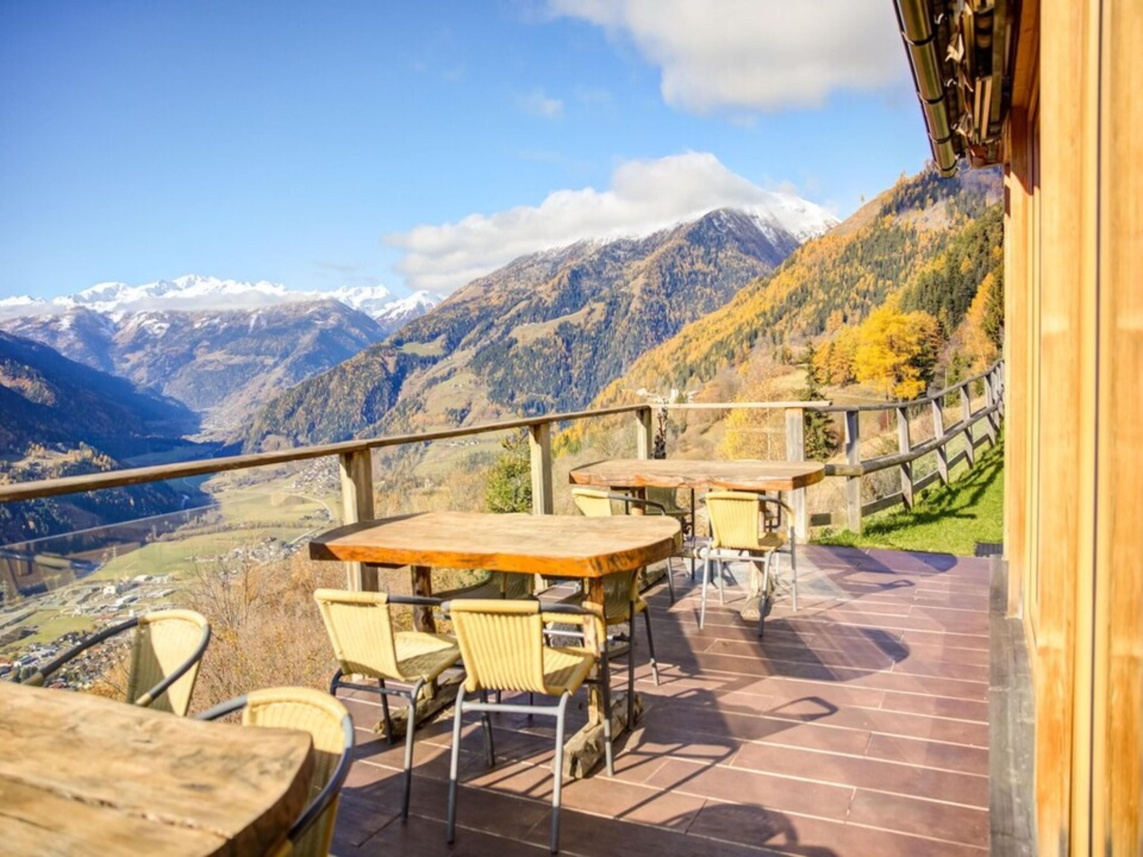 Une terrasse extérieure avec des tables et des chaises en bois, offrant une vue panoramique sur les montagnes et une vallée.