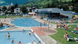Un espace piscine avec, en arrière-plan, un bâtiment portant l’inscription « ERLEBNISBAD », le tout entouré d’une végétation luxuriante et de montagnes.