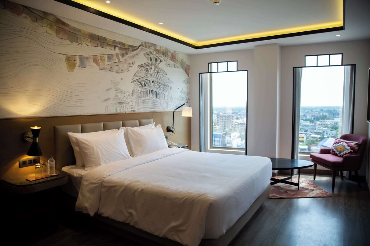 A modern hotel room with a large bed, a nightstand, a desk, a chair, and large windows offering a city view.