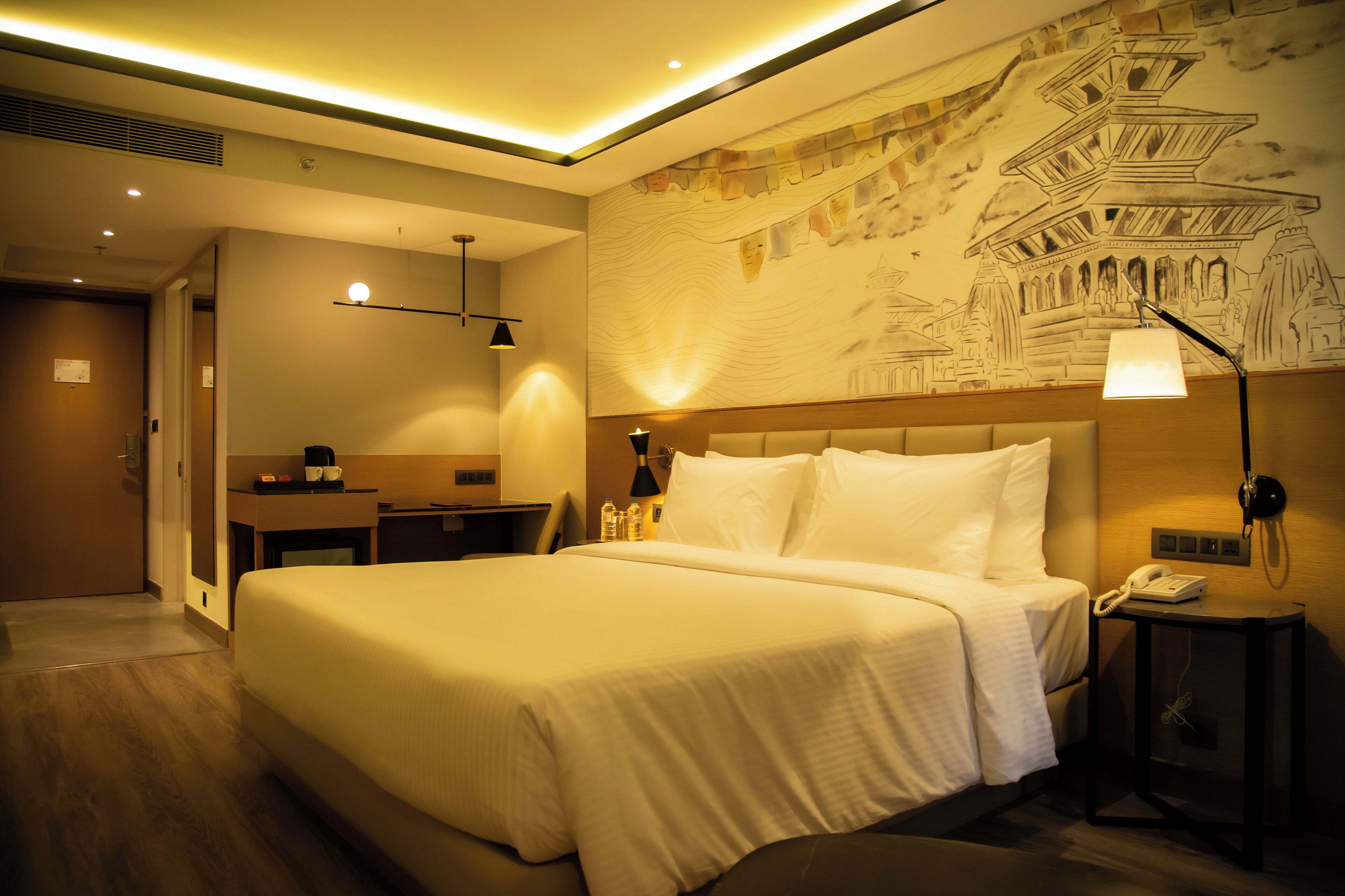 Quarto de hotel com uma cama grande, mesa de cabeceira, abajur e um mural na parede.