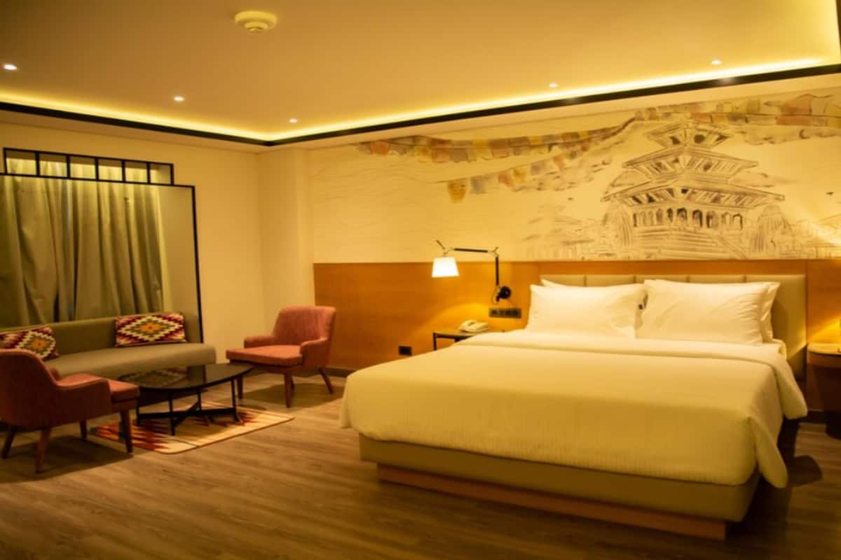 A hotel room with a large bed, a sofa, a chair, a desk, and a nightstand with a lamp.