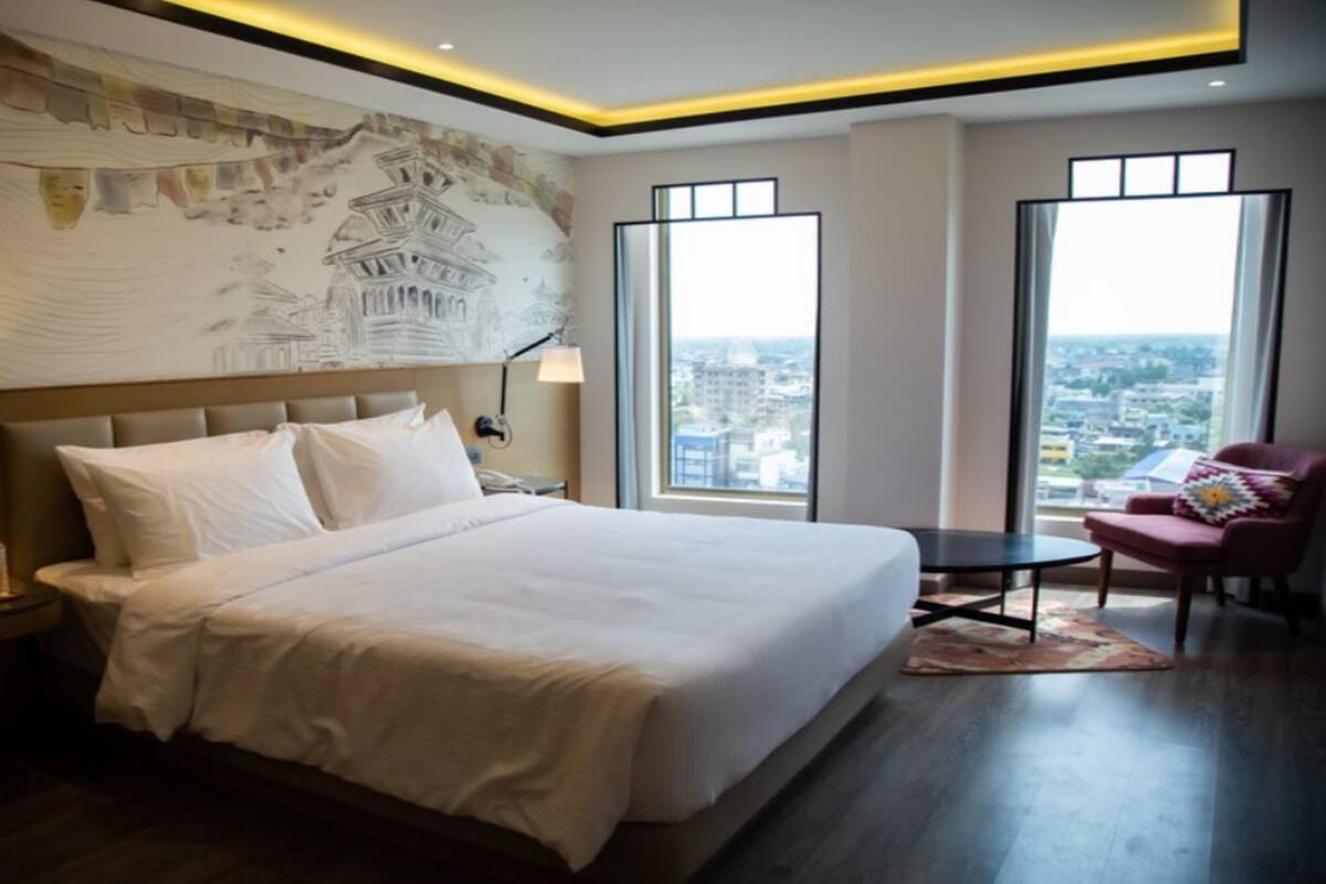A modern hotel room with a large bed, a nightstand, a lamp, a chair, and a large window offering a city view.