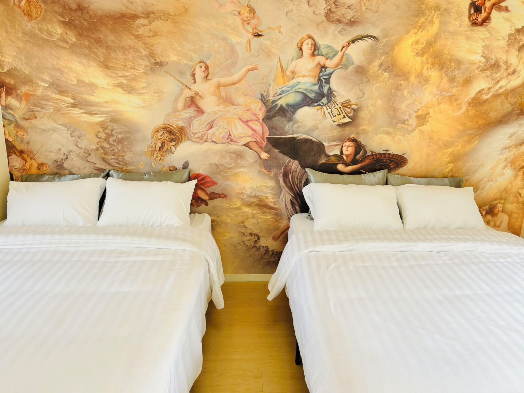 A hotel room with two beds, a mural of mythological figures on the wall, and a wooden floor.