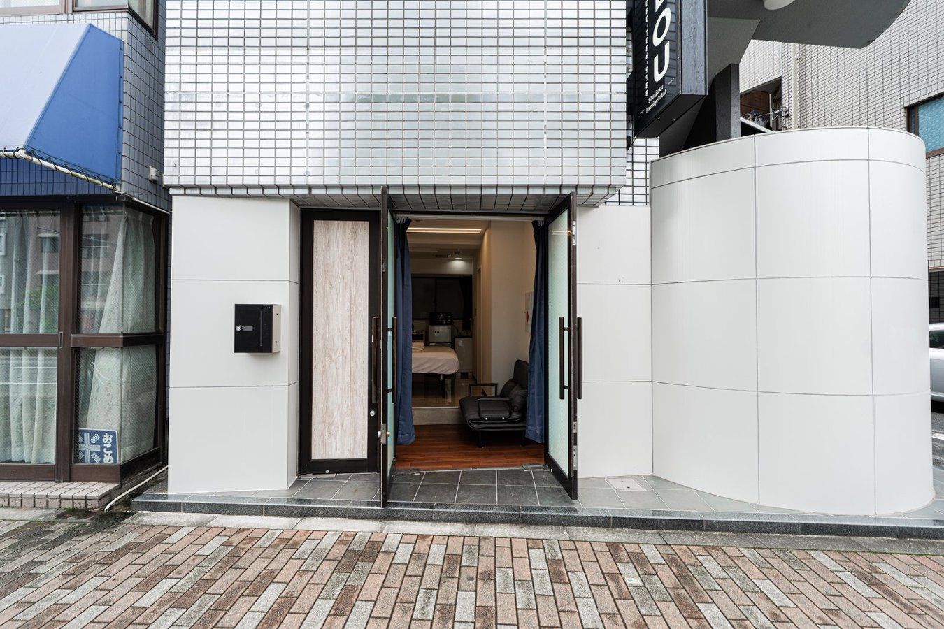 A hotel entrance with a tiled facade, a visible interior with a bed and a desk, and a sign with Japanese characters.